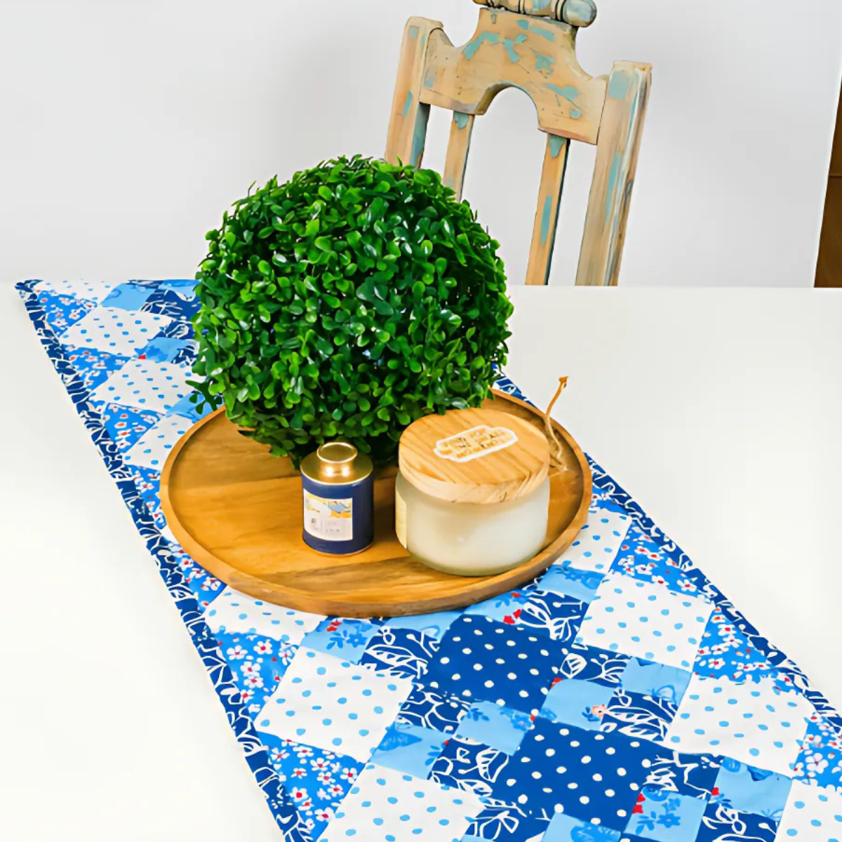 A wooden tray with a round faux plant, candle, and small jar rests on a white table adorned with the Table Runner Downloadable PDF Quilt Pattern; a distressed wooden chair is visible in the background.