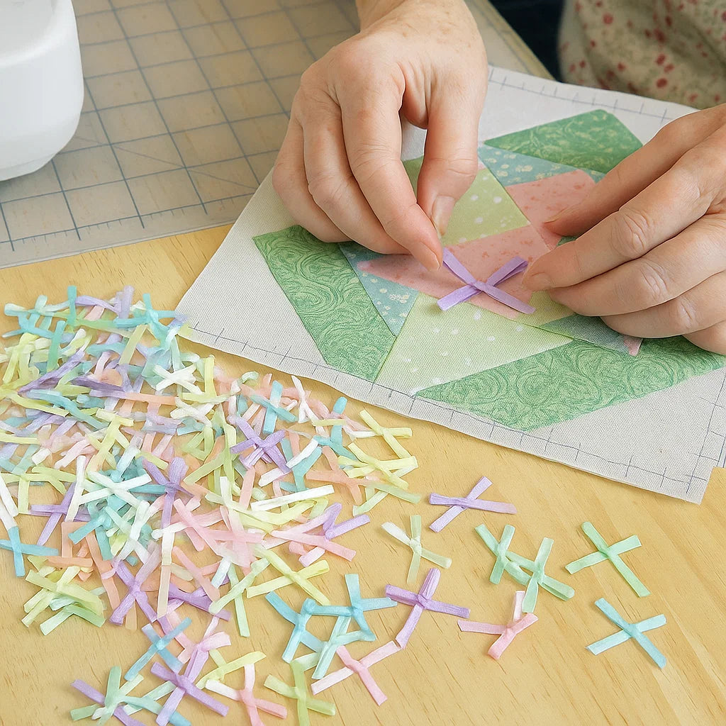 Hands arranging pastel fabric pieces and the Pastel Satin Ribbon Set on a quilt block, surrounded by matching fabric, sewing tools, and a partially visible sewing machine.