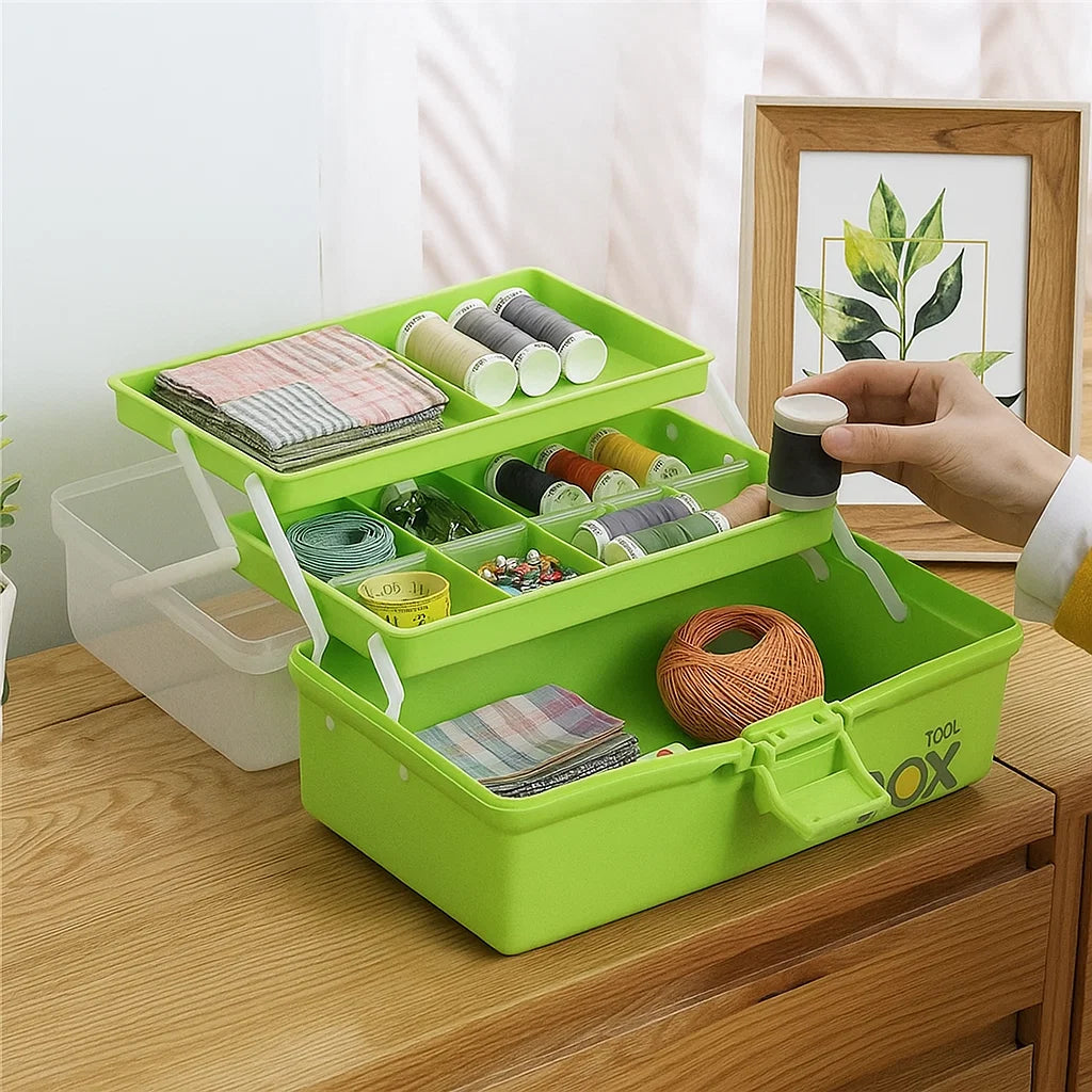 The Three-Layer Multipurpose Organizer Box in green, with compartments holding spools of thread, needles, fabric squares, and quilting tools, sits on a wooden table while a hand holds a spool of black and white thread.