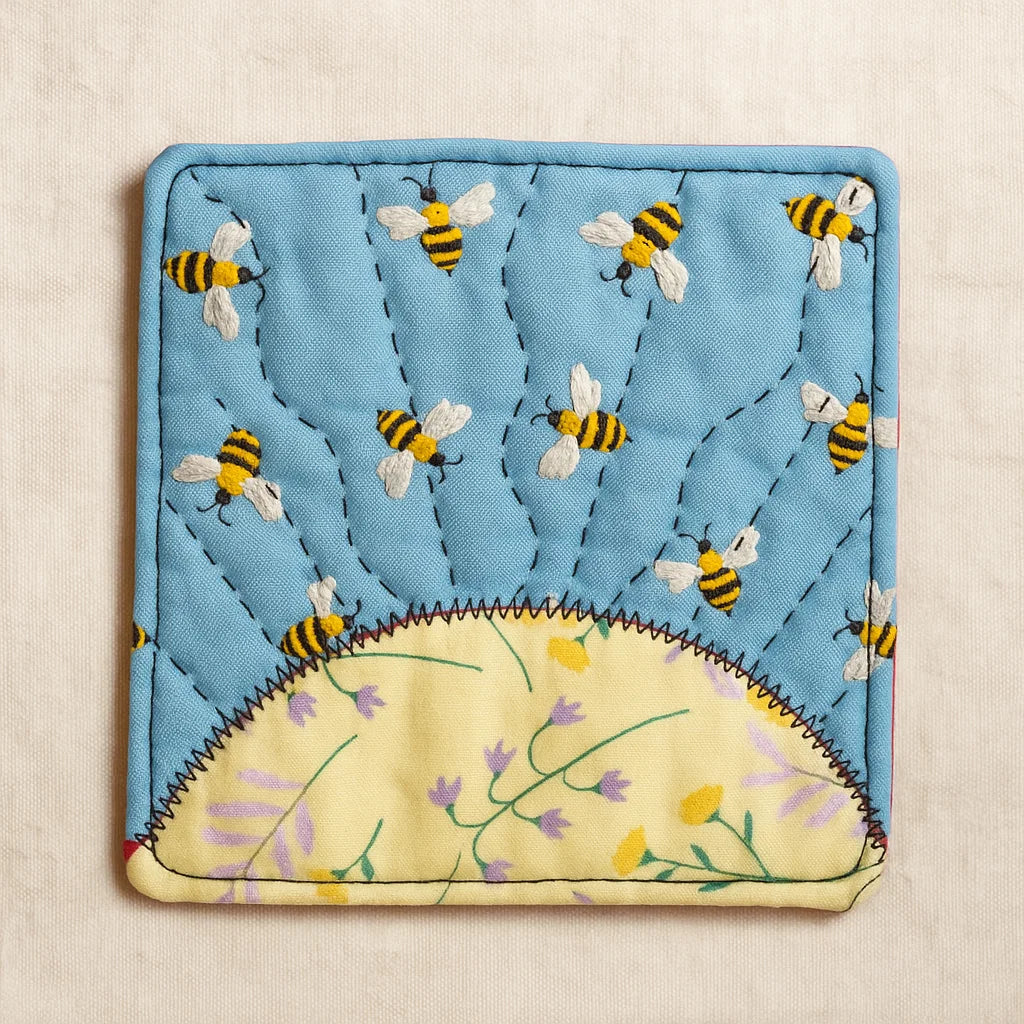 A square blue fabric potholder with bees and a yellow semi-circle with flowers is inspired by the Sunrise Coaster Downloadable PDF Quilt Pattern—ideal for anyone wanting a charming beginner quilt project.