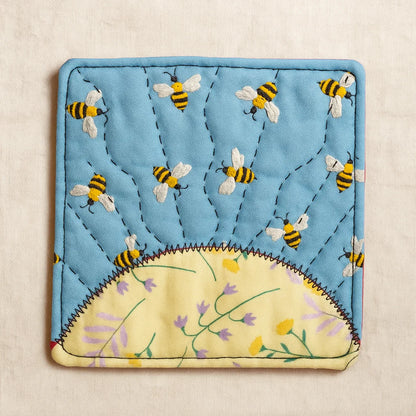A square blue fabric potholder with bees and a yellow semi-circle with flowers is inspired by the Sunrise Coaster Downloadable PDF Quilt Pattern—ideal for anyone wanting a charming beginner quilt project.