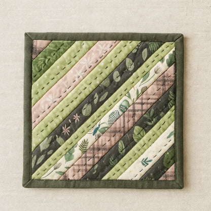 A square fabric coaster with diagonal quilted strips in green, pink, and cream botanical prints highlights the String Pieced Pot Holder Downloadable PDF Quilt Pattern—ideal inspiration for intermediate quilters.