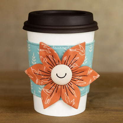 A white disposable coffee cup with a black lid, wrapped in a blue cozy made using the Flower Cup Sleeve Downloadable PDF Quilt Pattern, featuring a smiling orange fabric flower.