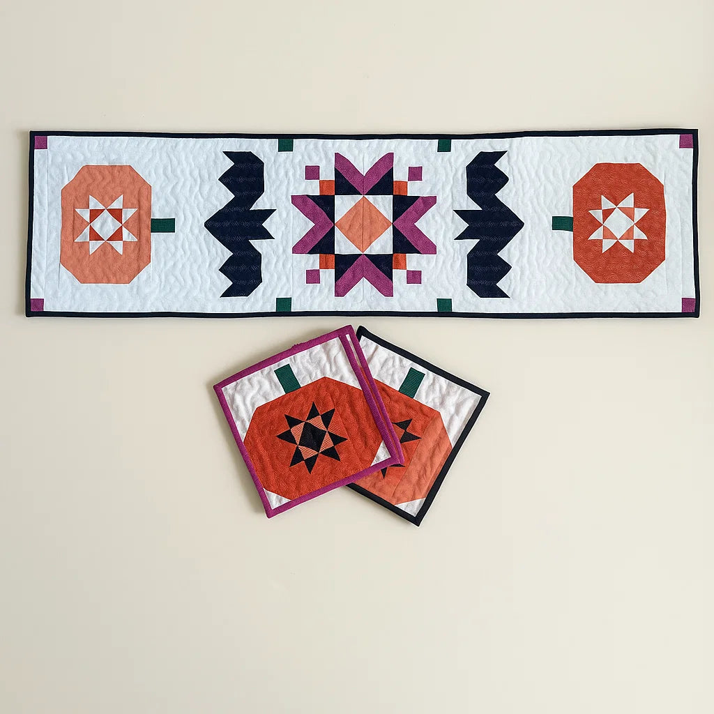 The Batty Harvest Downloadable PDF Quilt Pattern features a quilted table runner with geometric pumpkin and star designs, plus matching pumpkin coasters—perfect fall decor for your table on a light background.