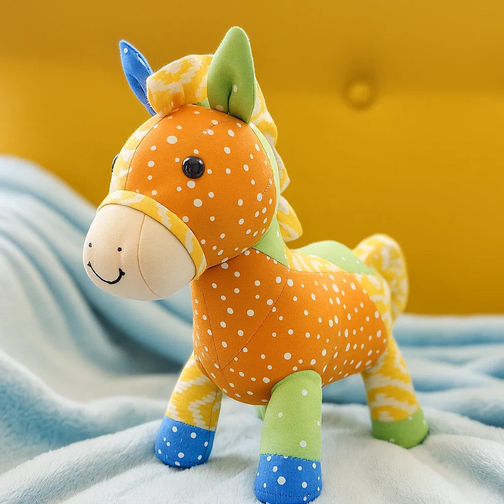 A colorful stuffed toy horse, inspired by the Sunny the Stallion Downloadable PDF Quilt Pattern, features orange, green, and blue patchwork and stands on a light blue blanket with a yellow cushion in the background.