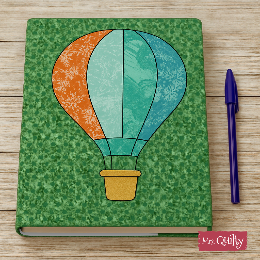 A Book Cover Downloadable PDF Quilt Pattern with a hot air balloon design is shown on a green notebook beside a blue pen, all resting on a wooden surface.