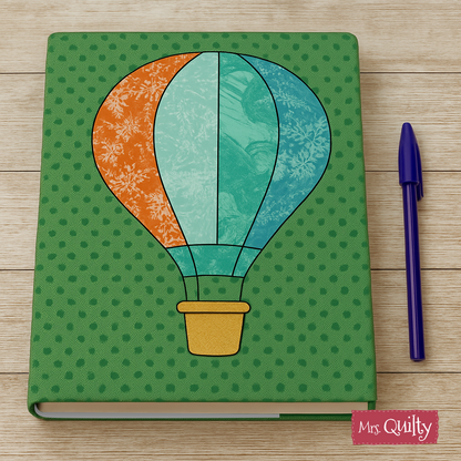 A Book Cover Downloadable PDF Quilt Pattern with a hot air balloon design is shown on a green notebook beside a blue pen, all resting on a wooden surface.
