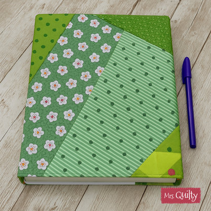 A digital PDF quilt pattern for a customizable book cover with green and floral patchwork designs is shown on a wooden surface beside a purple pen.