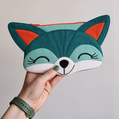 A hand holds the Freddie Fox Purse Downloadable PDF Quilt Pattern, a whimsical accessory in teal, white, and orange with a smiling face and closed eyes.