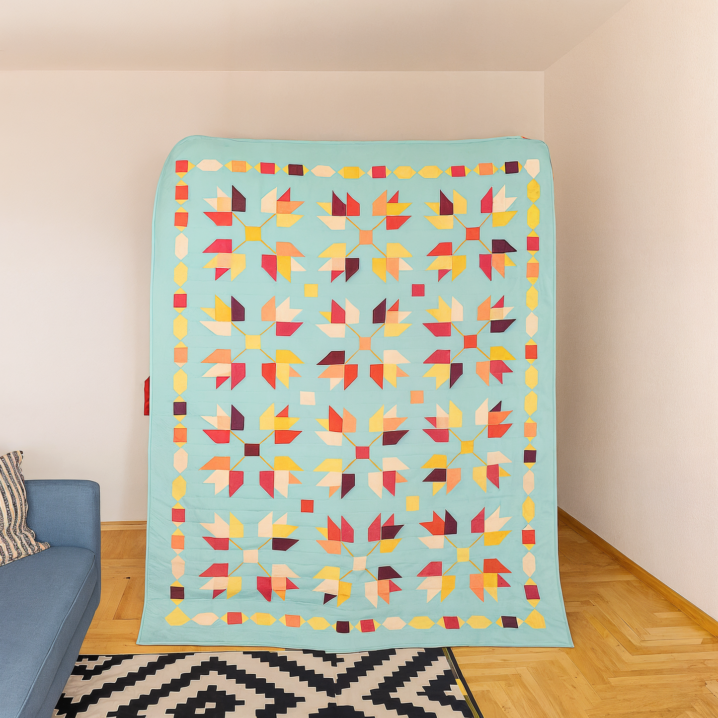 The Falling Autumn Downloadable PDF Quilt Pattern features a vibrant patchwork of geometric leaf designs in red, yellow, and orange on light blue, bringing cozy autumn vibes to your living room decor.