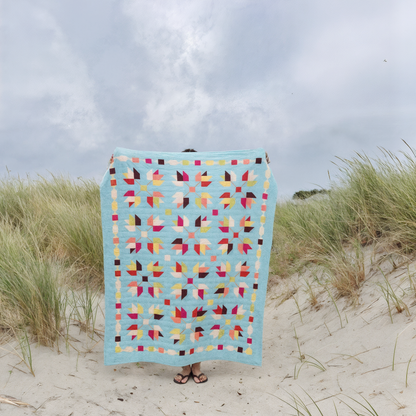 Standing on a sandy beach with tall grass, a person holds up the Falling Autumn Downloadable PDF Quilt Pattern, showcasing its colorful autumn design while their face and torso are hidden.
