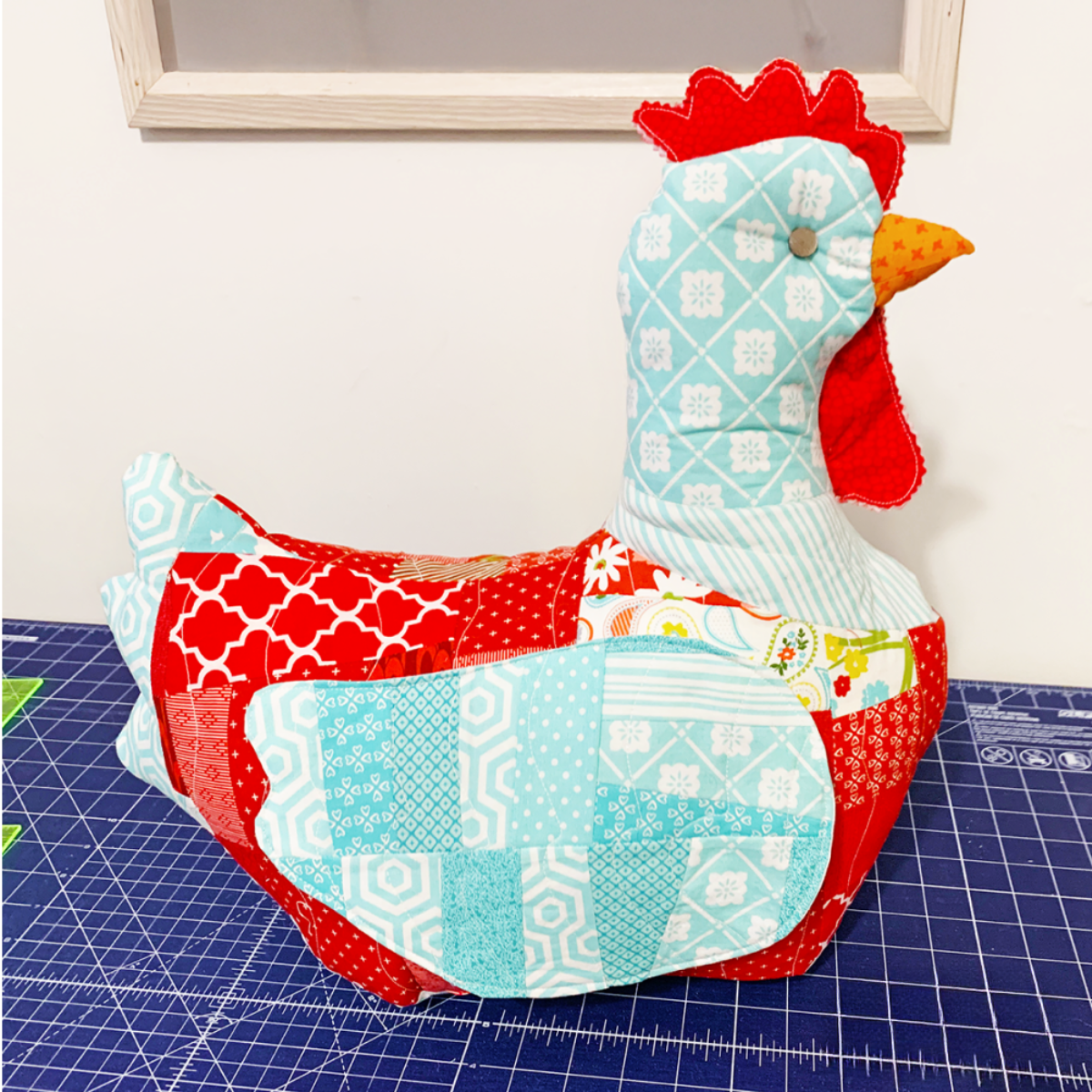 A chicken made from the Mrs. Cluckworth Downloadable PDF Quilt Pattern sits on a cutting mat in a craft room with red, blue, and white decor.