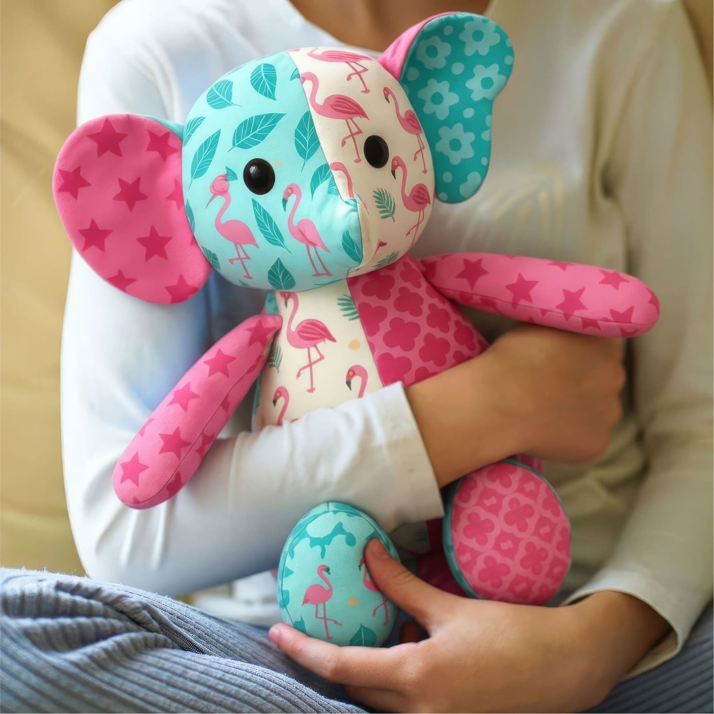 A person in a white top holds a colorful handmade stuffed elephant with mixed patterns—flamingos, stars, leaves, and flowers—in pink, teal, and cream. Find similar ideas in the Mrs. Quilty Plushies – Bundle of 14 Quilted Patterns.