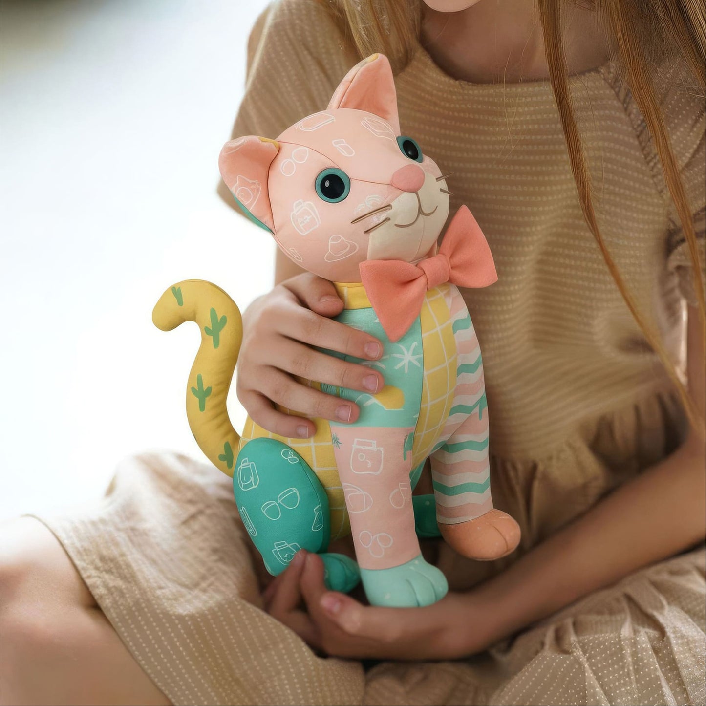 A child in a beige dress holds a pastel Mrs. Quilty Plushie cat from the Bundle of 14 Quilted Patterns, featuring a pink bow tie and colorful quilted designs.