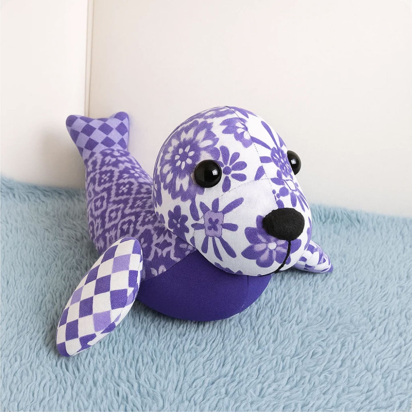 The Mrs. Quilty Plushies – Bundle of 14 Quilted Patterns features a plush seal toy with floral, checked, and solid purple fabrics resting on a blue textured surface against a light wall.