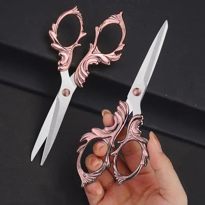 A person holds Embroidery Scissors featuring intricate rose gold handles and stainless steel blades, while another identical pair rests on a black surface, highlighting their vintage elegance.